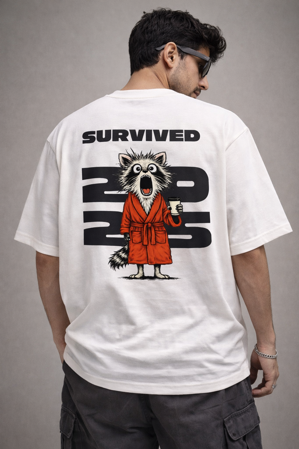 A model from the apparel brand Absolutely Nothing wearing a white oversized t-shirt with a graphic design and text on the back that says "Survived 2025", standing against a gray background.